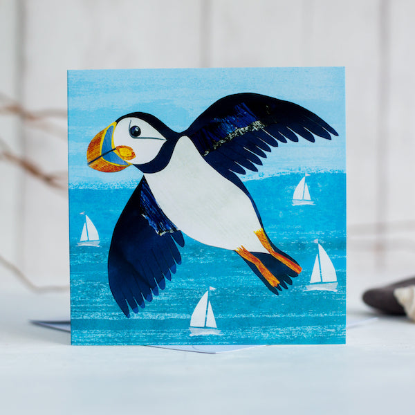 A greetings card showing c colourful  puffin flying over the turquoise sea. White sailing boats bob on the water below.