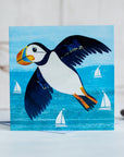 A greetings card showing c colourful  puffin flying over the turquoise sea. White sailing boats bob on the water below.