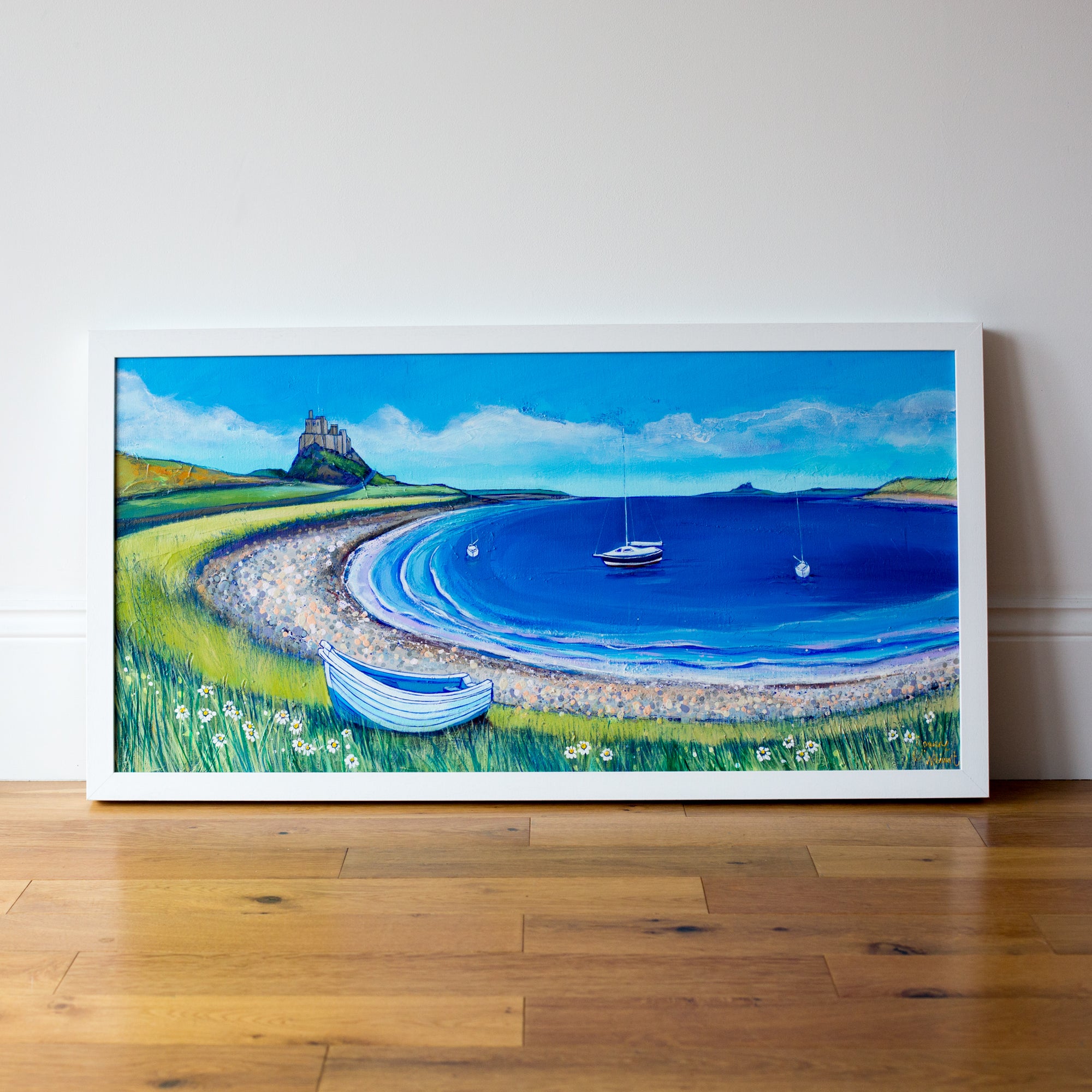 A coastal landscape painting in a white wood frame  featuring Lindisfarne Castle iwith the bay circling in the forground the sea and sky ar ebrigh blue with a white coble boat resting on the shore. 