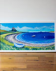 A coastal landscape painting in a white wood frame  featuring Lindisfarne Castle iwith the bay circling in the forground the sea and sky ar ebrigh blue with a white coble boat resting on the shore. 