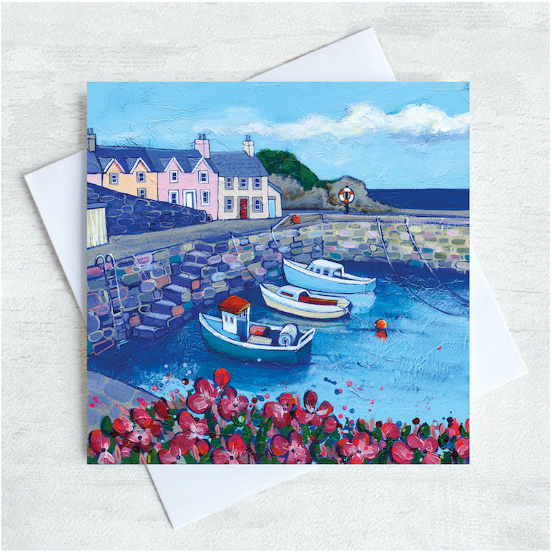 Three fishing boats sit in the harbour with some pasetel coloured houses above the harbour wall. Pink flowers adorn the foreground. 