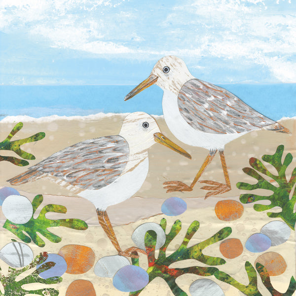 Seaside scene with a pale blue sea and blue cloudy sky. The sand is littered with green seaweed and red, blue and grey rocks but the stars of the show are two Sanderling birds pottering along the shore line. These birds have a white head and stomach and beige/brown textured wings, beaks and legs. 