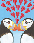 Two puffins stand beak to beak with multiple textured red love hearts between them. The puffins have their characteristic markings of bright orange and yellow beaks with white chests and faces. 