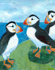 An art print of 3 cheeky puffins perched on a grassy ledge above the bright blue sea.