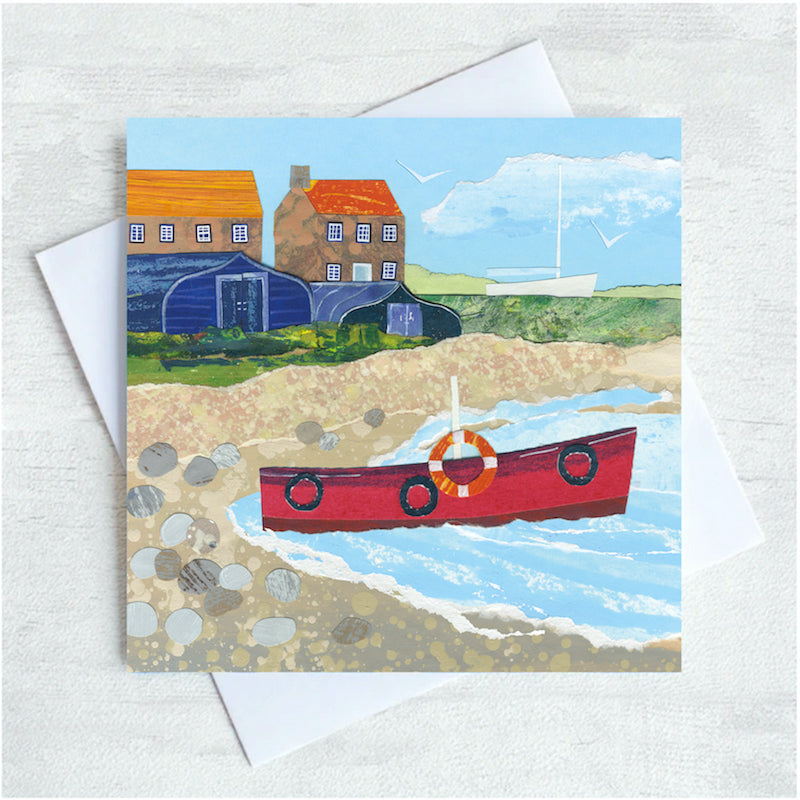 A seaside greetings card featuring a red fishing boat on a pebbled shoreline with upturned boats and cottages in the background. 