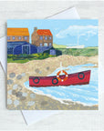 A seaside greetings card featuring a red fishing boat on a pebbled shoreline with upturned boats and cottages in the background. 