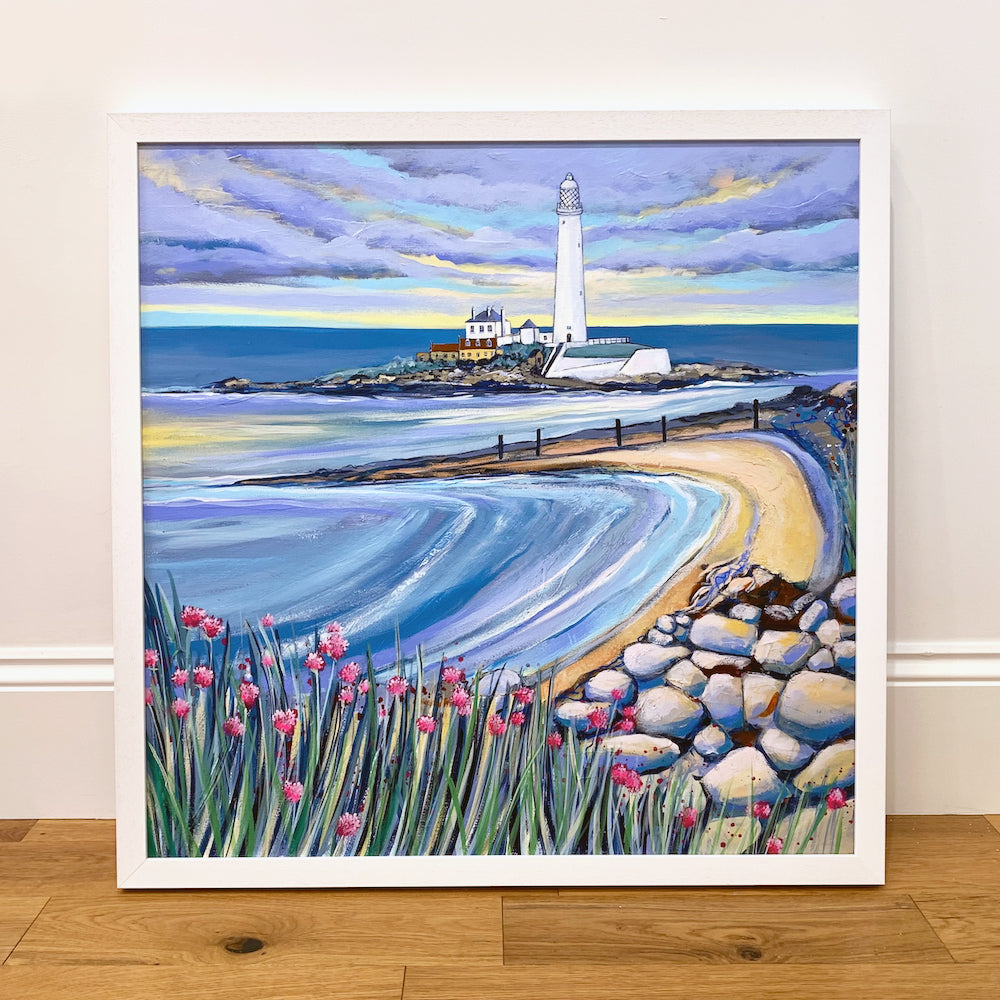A painting in a white farne stands on a wooden floor against  a white wall. THe painting is of a white lighthouse with a small beach scene and pebbled shore. Swathes of sea pink flowers are displayed in the foreground.. 