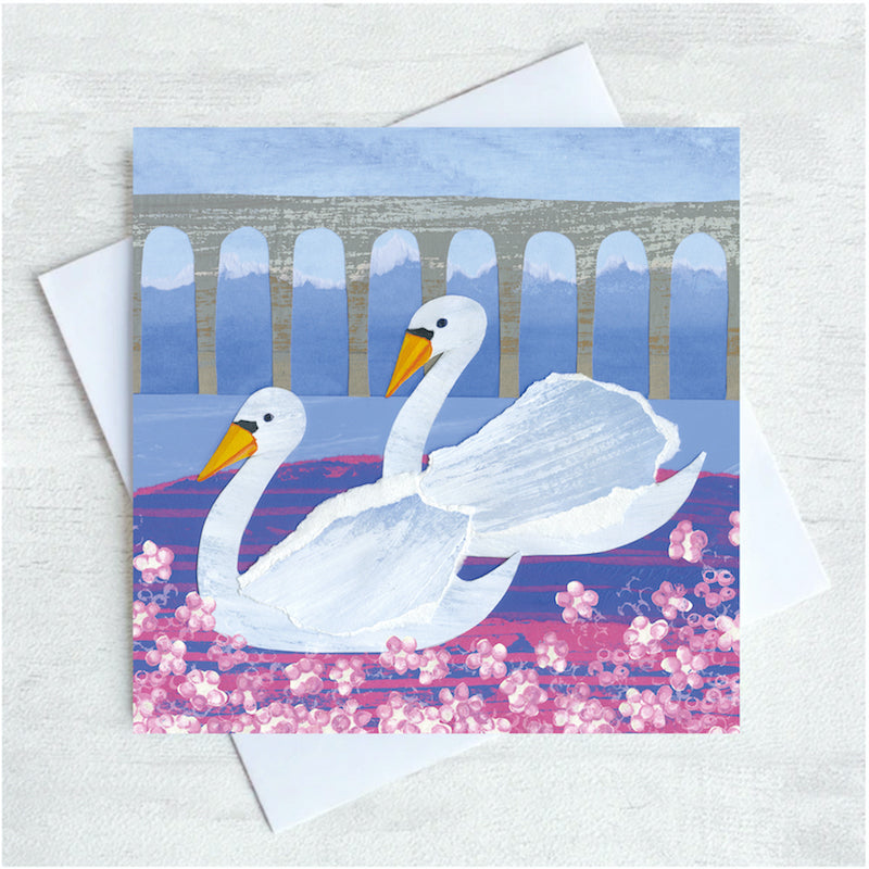 Two beautiful swans glide on a river with an arched bridge overhead. Pink flowers pepper the foreground.  