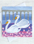 Two beautiful swans glide on a river with an arched bridge overhead. Pink flowers pepper the foreground.  