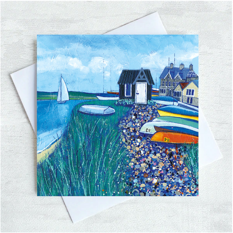 A cute little blck hut sits on the shore alongside the river with a sailing boat passing by. Brightly coloured boats are pile up alongside a pebbled path. 