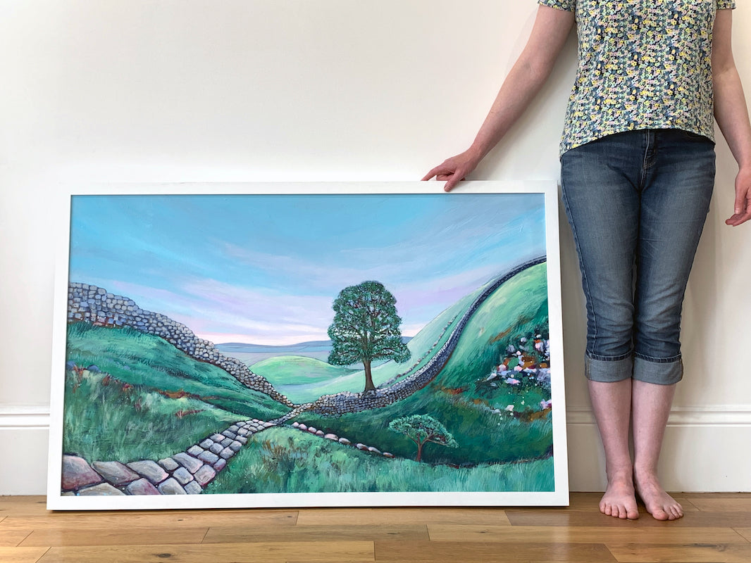 Artist Joanne wisharts legs stand next to a large painting of Sycamore Gap. 