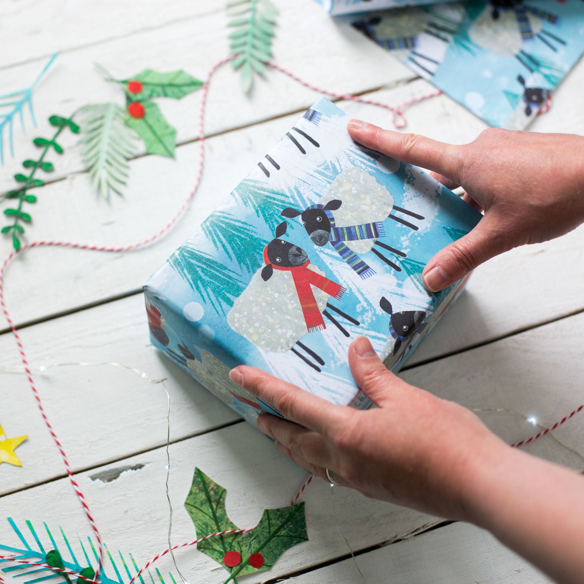 In this image two hands are placed on top of a cuboid parcel wrapped in the winter woolies gift wrap. The gift wrap section visible shows two black-headed sheep facing each other. One wears a red scarf while the other wears a purple toned striped scarf. The background is blue with Christmas trees and snow dispersed between the cheery characters.