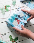 In this image two hands are placed on top of a cuboid parcel wrapped in the winter woolies gift wrap. The gift wrap section visible shows two black-headed sheep facing each other. One wears a red scarf while the other wears a purple toned striped scarf. The background is blue with Christmas trees and snow dispersed between the cheery characters.