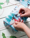 In this image two hands are busy tying a bow with white and red string on top of a parcel wrapped in the winter woolies gift wrap. The gift wrap section visible shows two black-headed sheep facing each other. One wears a red scarf while the other wears a purple toned striped scarf. The background is blue with Christmas trees and snow dispersed between more cheery sheep characters.