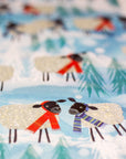 Close up view of the wrapping paper. In focus are two black-headed sheep facing each other. One wears a red scarf while the other wears a purple toned striped scarf. The background is blue with Christmas trees and snow dispersed between the cheery characters.